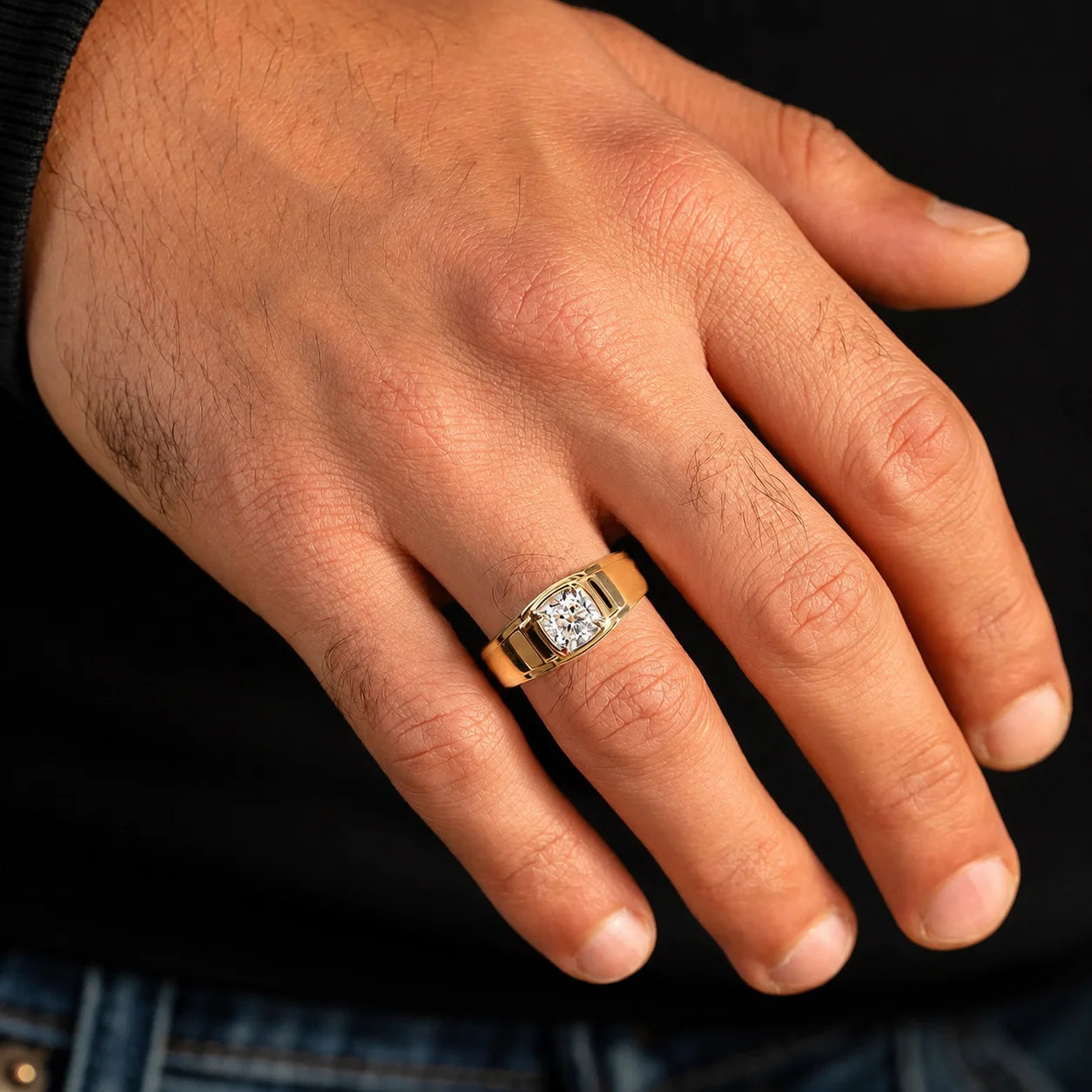 A close-up of a man's hand wearing a yellow gold ring with a square-cut diamond set in a bezel setting, against a dark background.