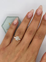 A close-up of a hand wearing a silver ring with an emerald-cut clear center stone and small side stones, with a pale blue jewelry box partially visible in the background.