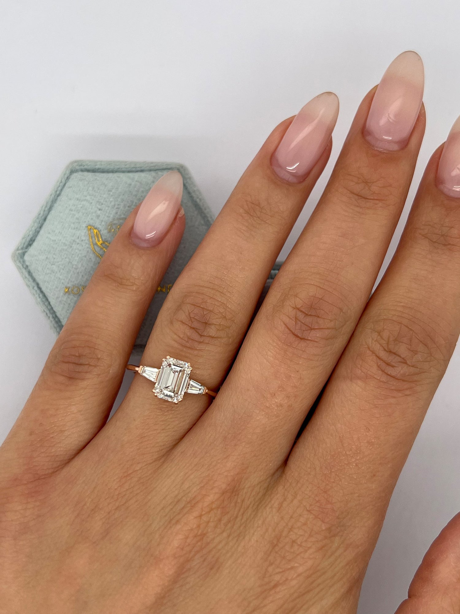 A close-up of a hand wearing a silver ring with an emerald-cut clear center stone and small side stones, with a pale blue jewelry box partially visible in the background.