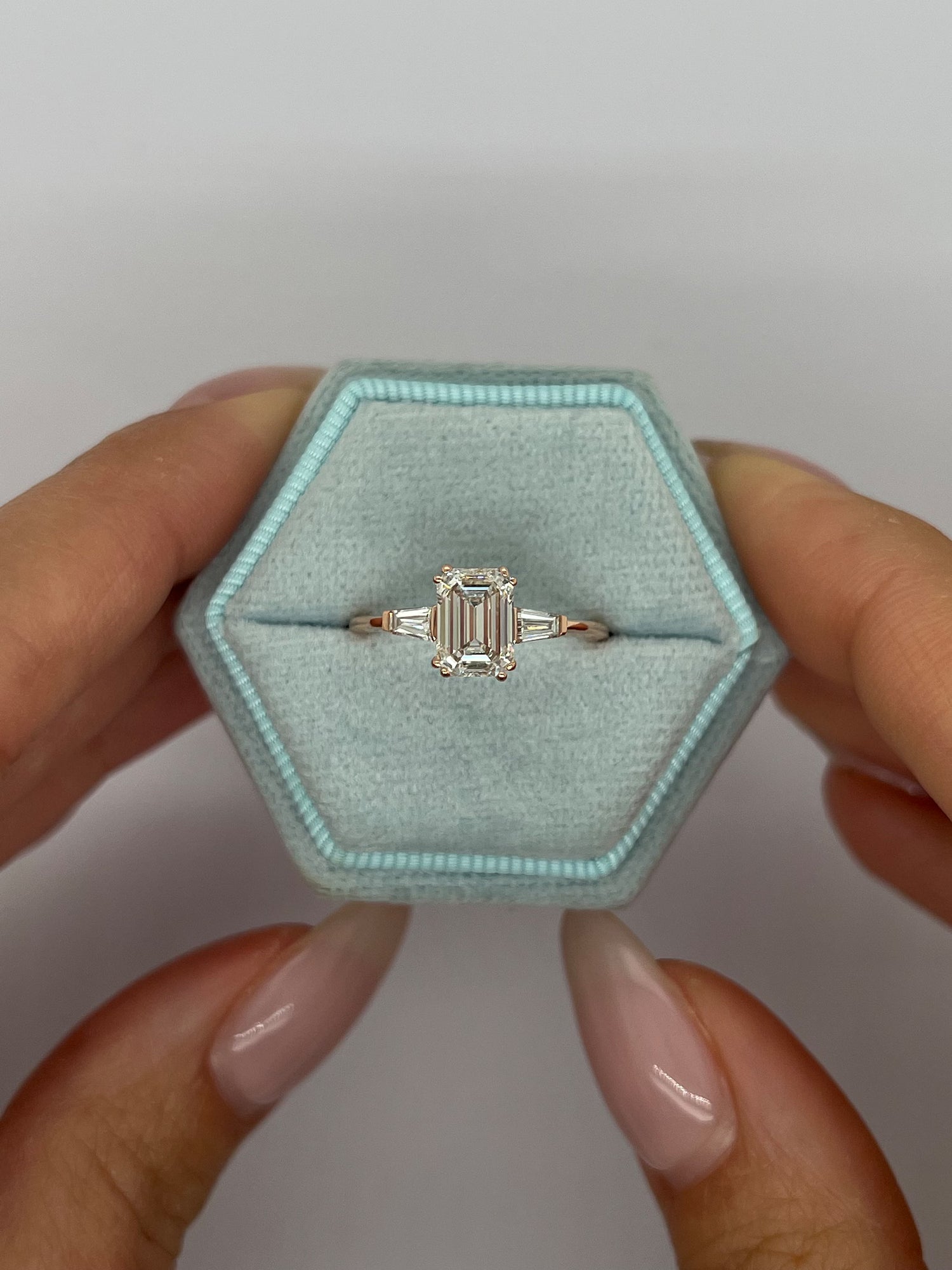 A silver emerald-cut ring held in a light blue hexagonal ring box, with a person's hands visible holding the box against a plain light gray background.