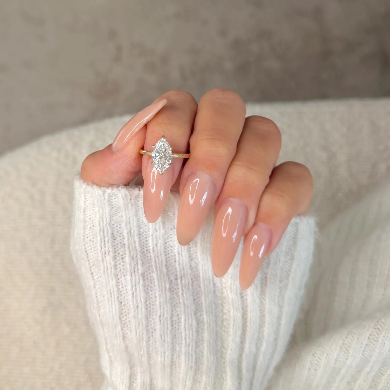 A close-up of a hand with long, glossy nude manicured nails wearing a gold ring with a marquise-cut clear gemstone, resting on a soft white ribbed knit sleeve.