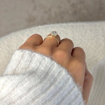 A close-up of a hand wearing a gold ring with a clear heart-shaped gemstone, set against a soft white knit sweater and a neutral beige background.