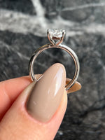 A close-up of a silver engagement ring with a clear round-cut gemstone held by a hand with beige manicured nails, set against a dark gray marble background.