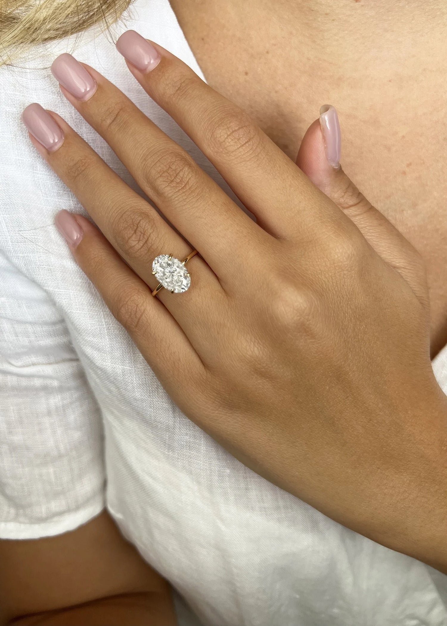 A close-up of a person's hand wearing a gold ring with a large oval-cut clear gemstone, resting on their chest against a white top.