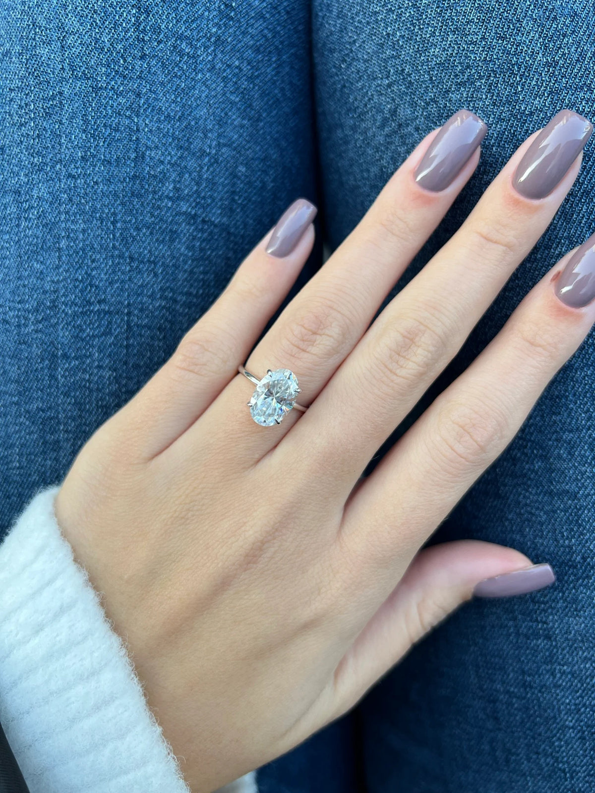 A close-up of a hand with mauve-colored nails wearing a silver ring with a large clear gemstone, resting against blue denim fabric.