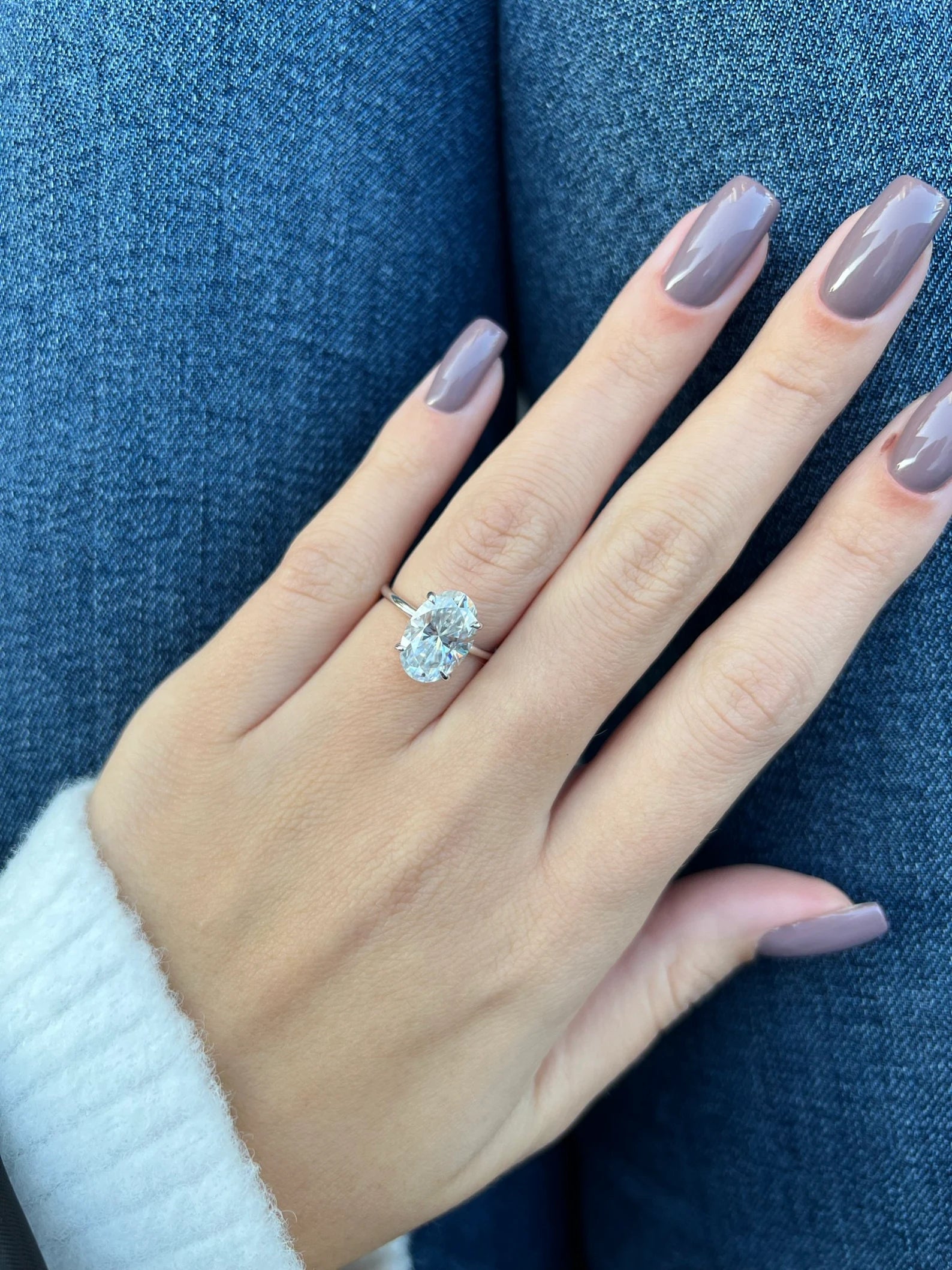 A close-up of a hand with mauve-colored nails wearing a silver ring with a large clear gemstone, resting against blue denim fabric.