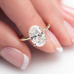 An oval-cut clear gemstone ring in a gold four-prong setting, shown worn on a finger with a soft-focus background.