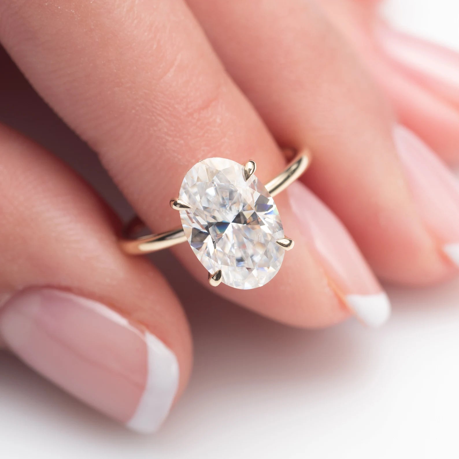 An oval-cut clear gemstone ring in a gold four-prong setting, shown worn on a finger with a soft-focus background.