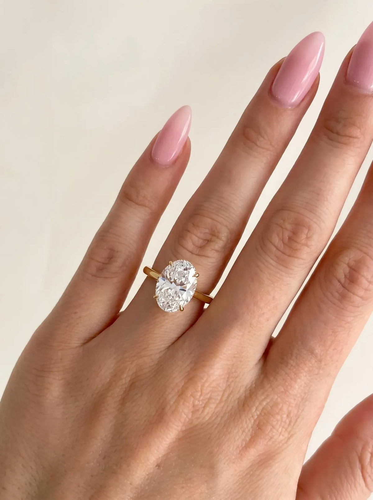 A close-up of a hand with long pink manicured nails wearing a yellow gold ring with a large oval-cut clear gemstone, set against a plain light beige background.