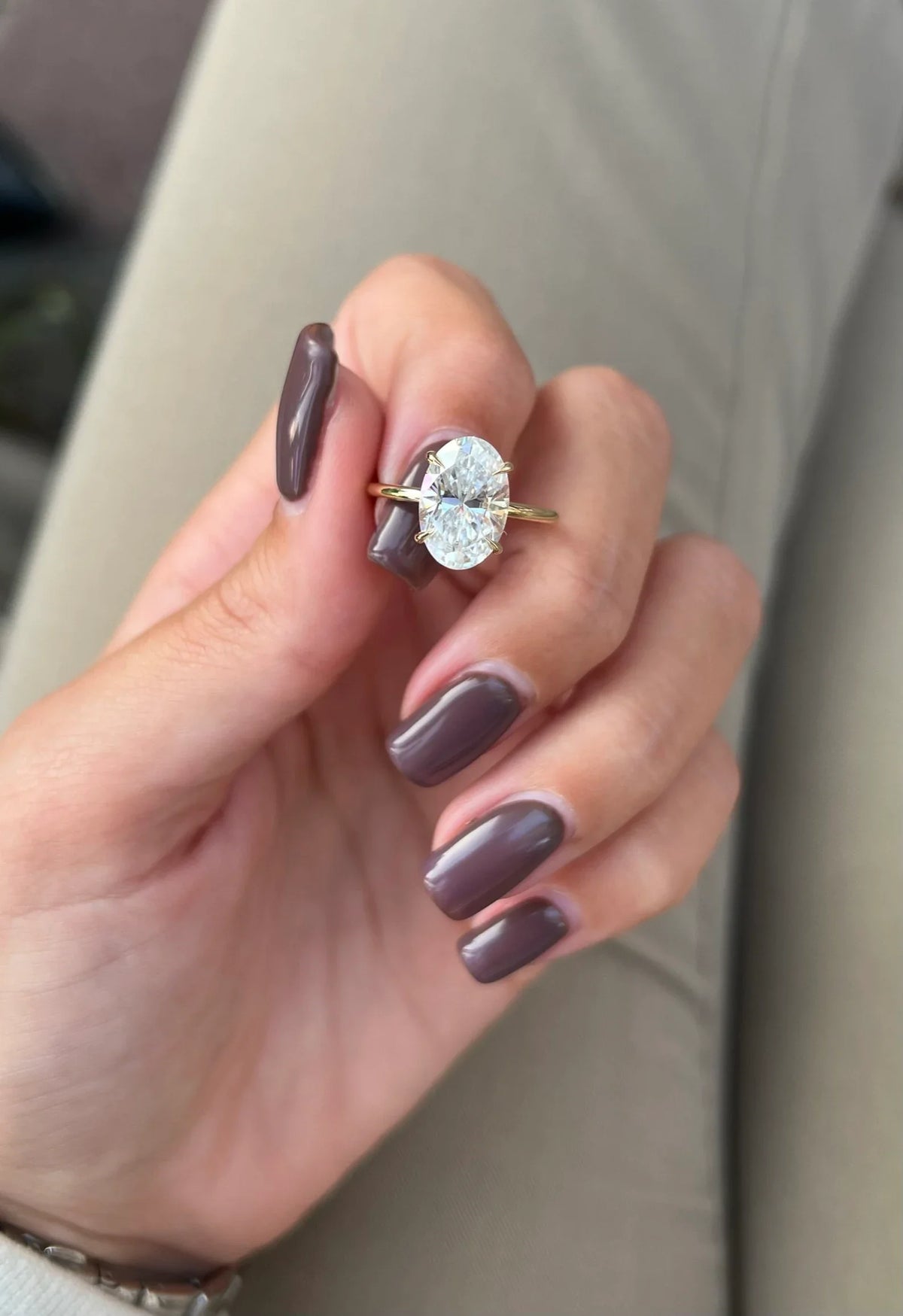 A close-up of a hand with mauve-colored nails wearing a gold ring with a large clear oval gemstone, set against a neutral beige background.