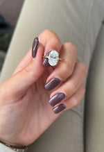 A close-up of a hand with mauve-colored nails wearing a gold ring with a large clear oval gemstone, set against a neutral beige background.