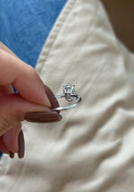 A close-up of a hand with brown manicured nails holding a silver ring with a small clear gemstone in a prong setting, against a background of blue denim and beige fabric.