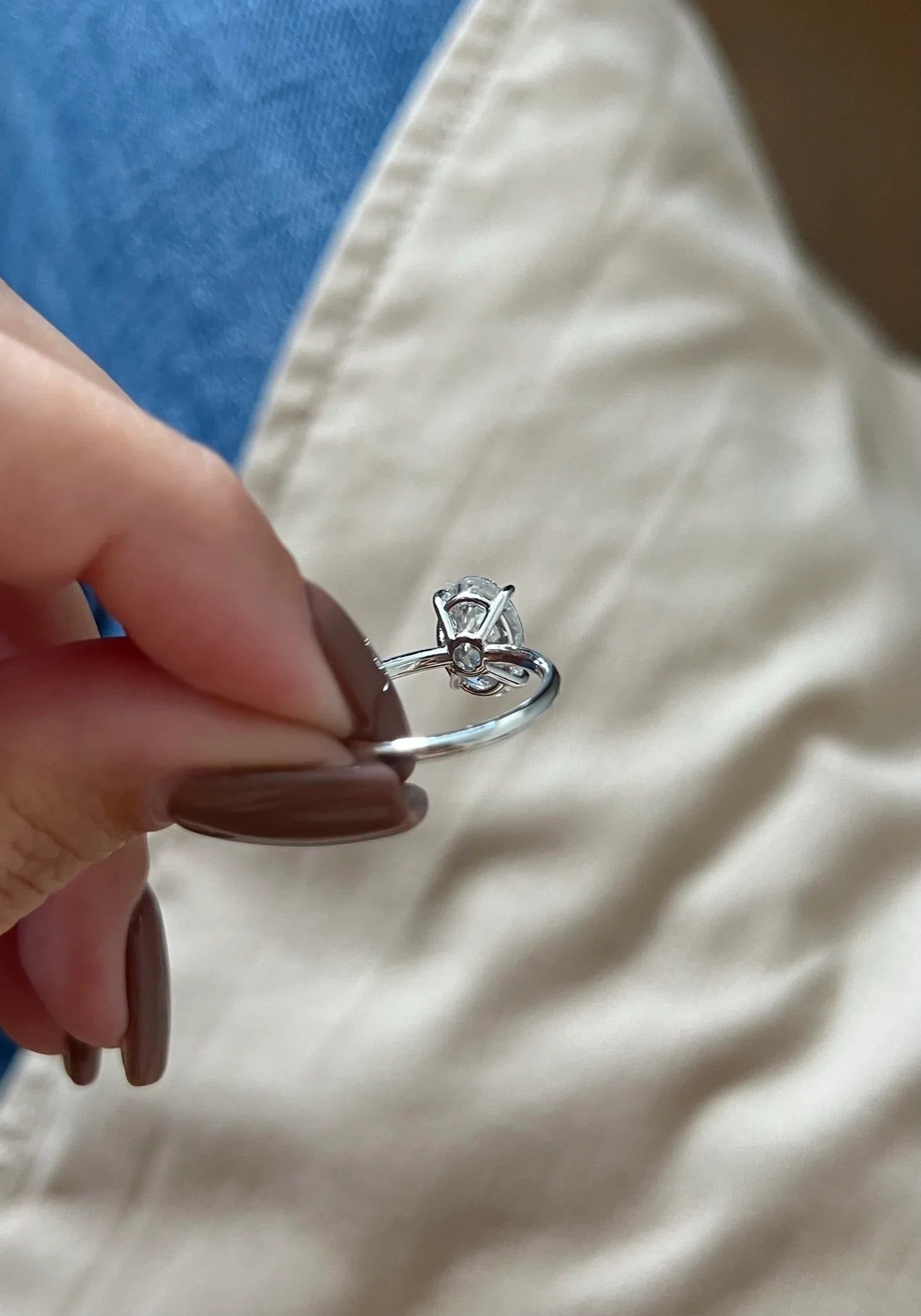 A close-up of a hand with brown manicured nails holding a silver ring with a small clear gemstone in a prong setting, against a background of blue denim and beige fabric.