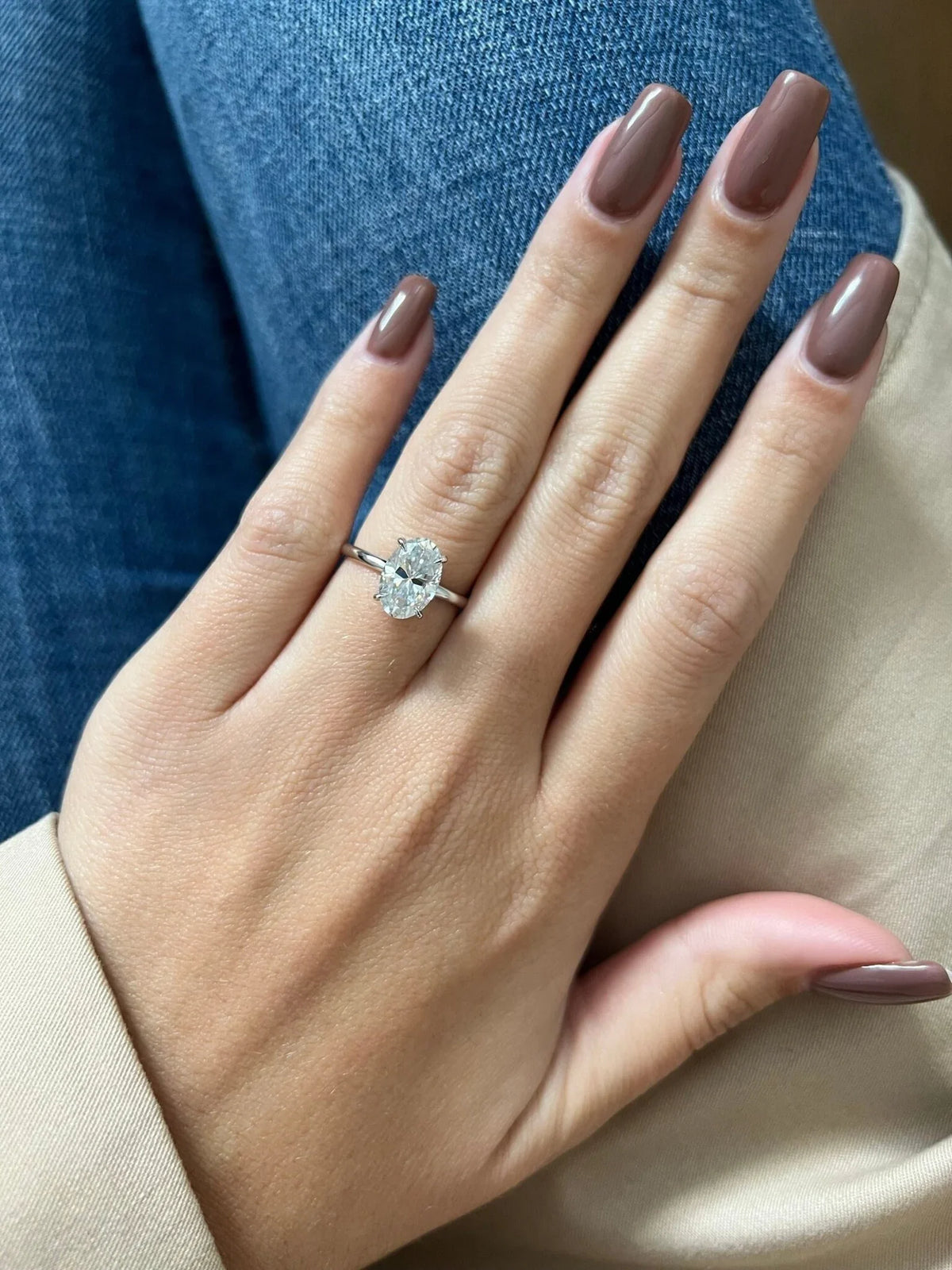 A close-up of a hand with mauve-brown manicured nails wearing a silver-toned ring with a clear oval-cut center stone, resting against blue denim and a beige fabric background.