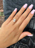 A close-up of a hand with light pink manicured nails wearing a rose gold ring with a round-cut clear gemstone, resting on dark gray denim fabric.