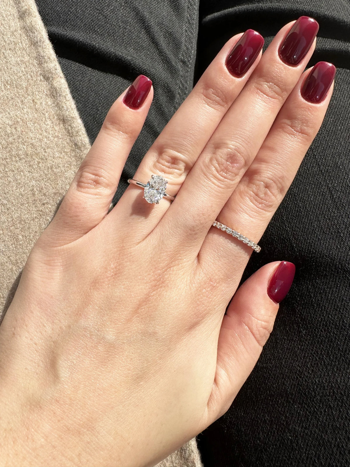A close-up of a hand with dark red manicured nails wearing a silver diamond engagement ring with a round center stone and a thin diamond band on another finger, set against dark clothing.