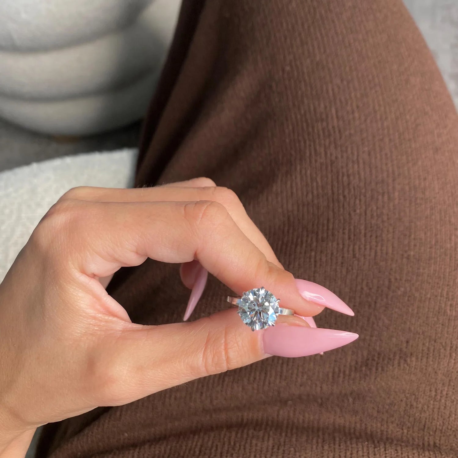 A close-up of a hand with long pink manicured nails holding a silver ring with a large round clear gemstone, set against brown knit clothing.