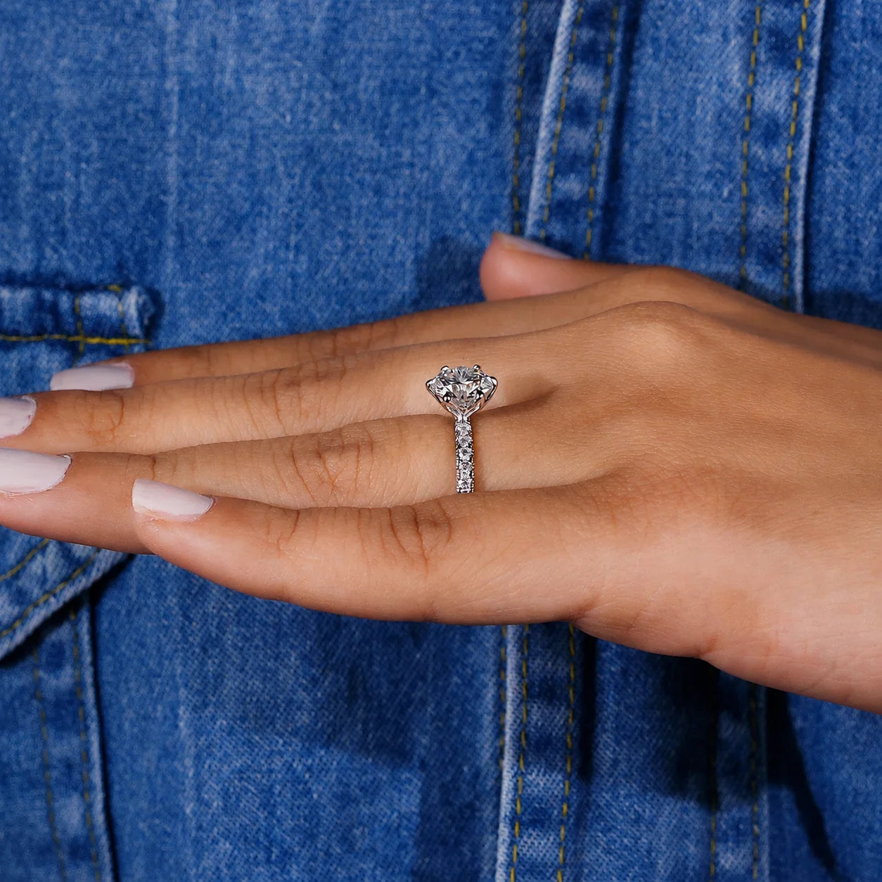 A close-up of a hand wearing a silver-toned diamond engagement ring, resting against a blue denim jacket.