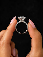 A silver-toned diamond engagement ring held between two hands with pink manicured nails against a solid black background.