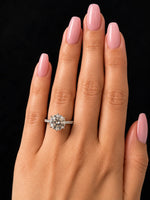 A close-up of a hand with long, neatly manicured pink nails wearing a round brilliant-cut diamond engagement ring on a black background.