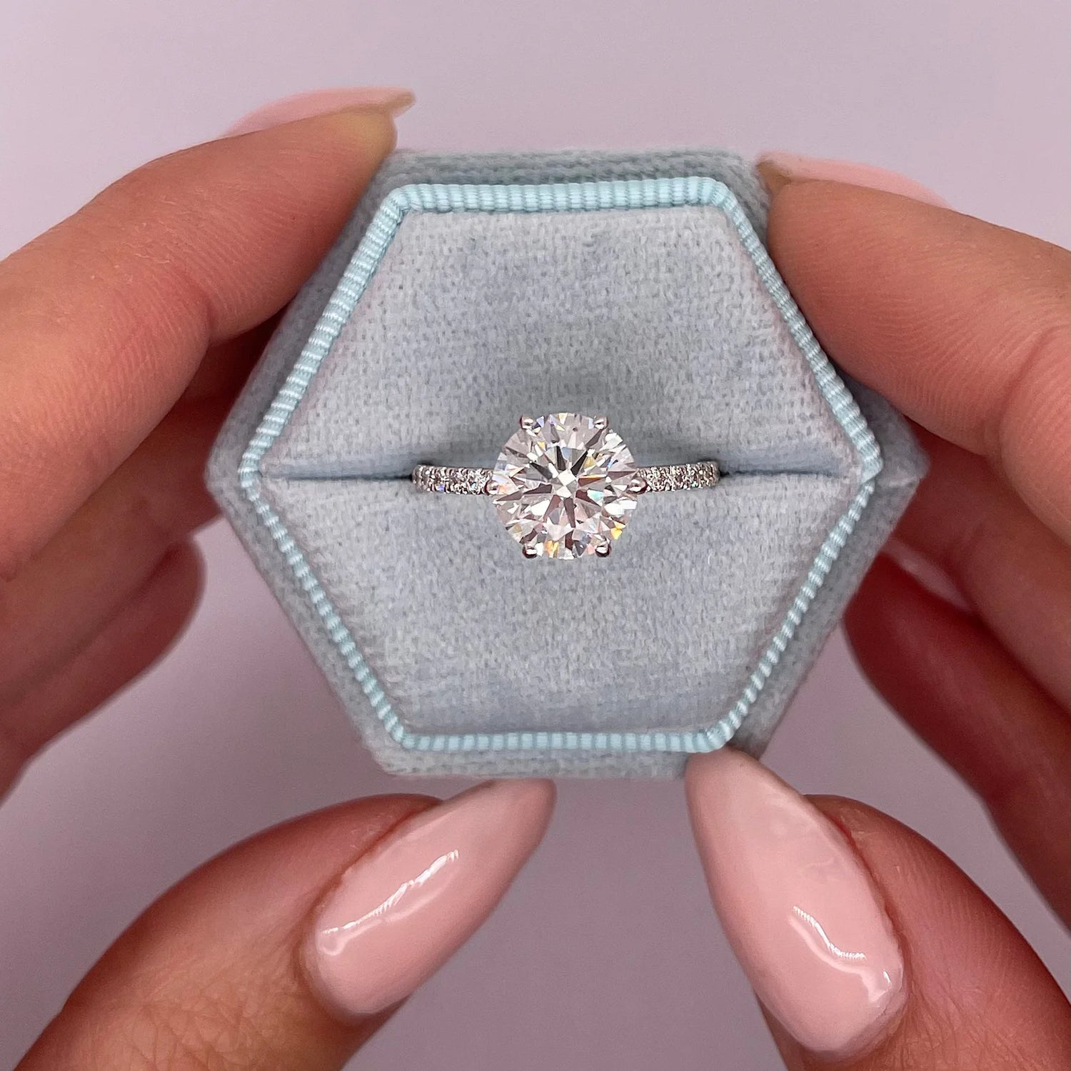 A round-cut diamond engagement ring with a silver-toned band, displayed in a hexagonal light blue velvet ring box held by two hands with pink manicured nails.