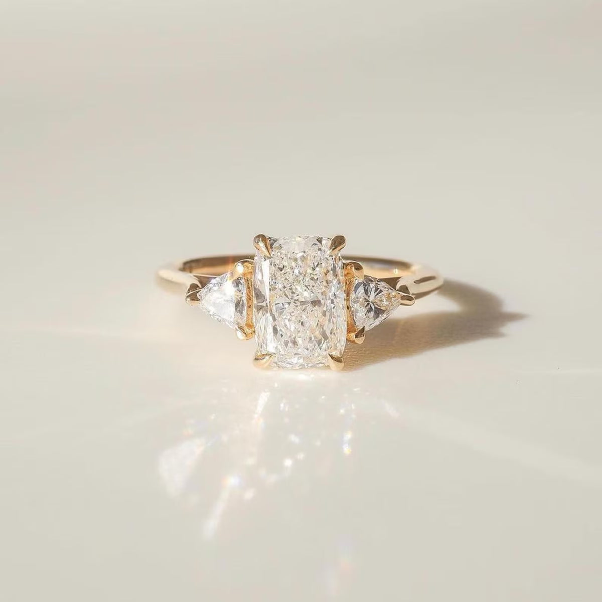 A three-stone diamond ring with a central rectangular radiant-cut diamond flanked by two smaller round-cut diamonds, set in a yellow gold band, photographed on a light beige reflective surface.