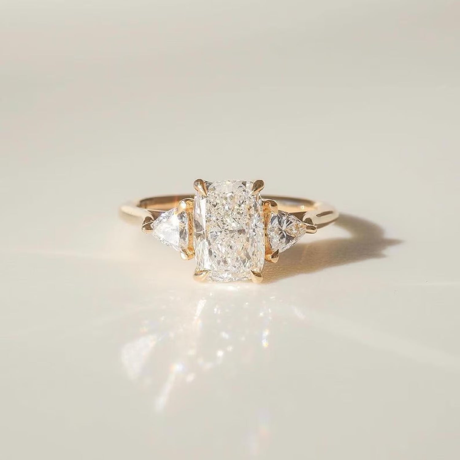 A three-stone diamond ring with a central rectangular radiant-cut diamond flanked by two smaller round-cut diamonds, set in a yellow gold band, photographed on a light beige reflective surface.